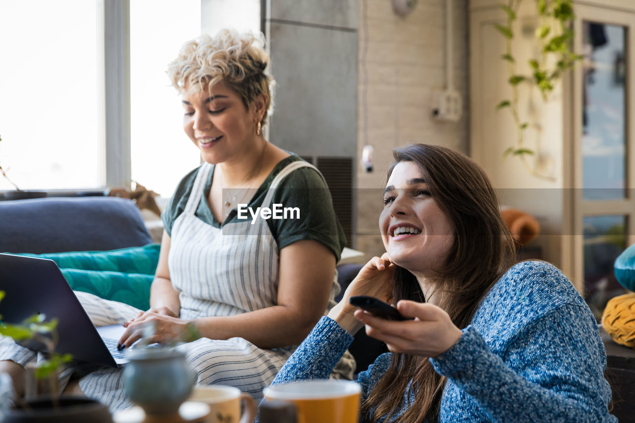 Happy young woman watching tv while friend using laptop at home