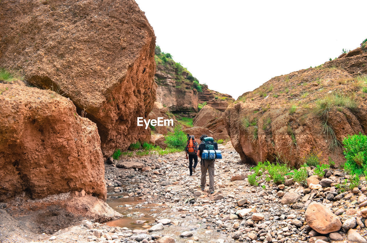 REAR VIEW OF MEN WALKING ON ROCKS AGAINST MOUNTAINS