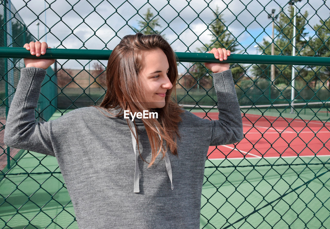 PORTRAIT OF BEAUTIFUL YOUNG WOMAN STANDING BY CHAINLINK FENCE IN A PARK