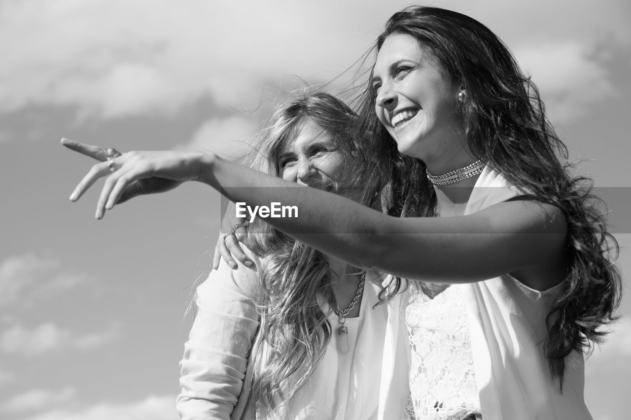 Cheerful young friends embracing while standing outdoors against sky