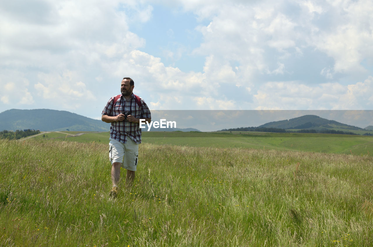 Man with a backpack and binoculars hiking through mountain meadow on a spring day.