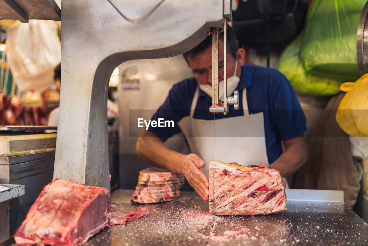 Mature man in apron and face mask using electric tabletop saw to slice piece of frozen meat during work in butchery