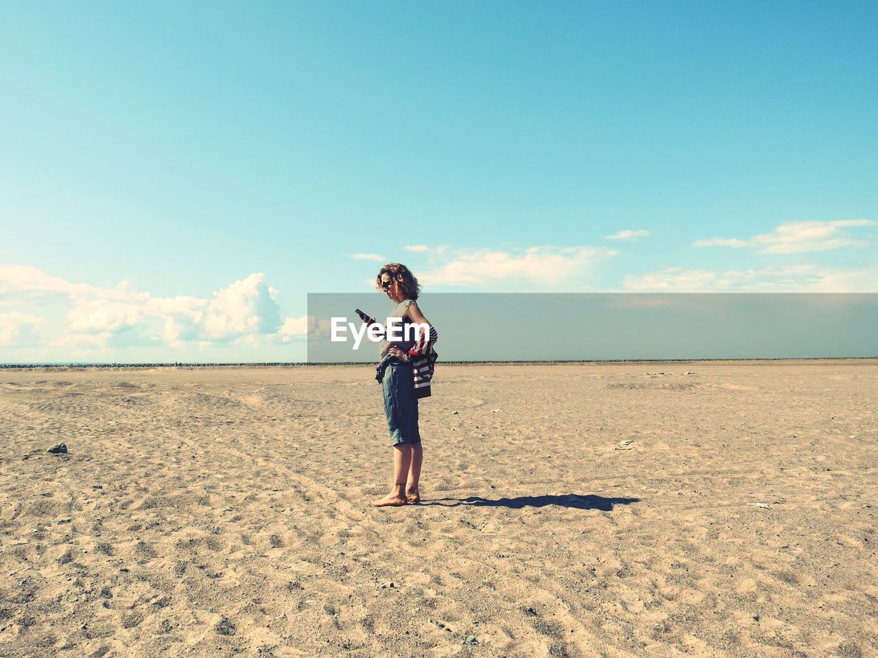 Woman using phone while standing on sand at desert against sky 