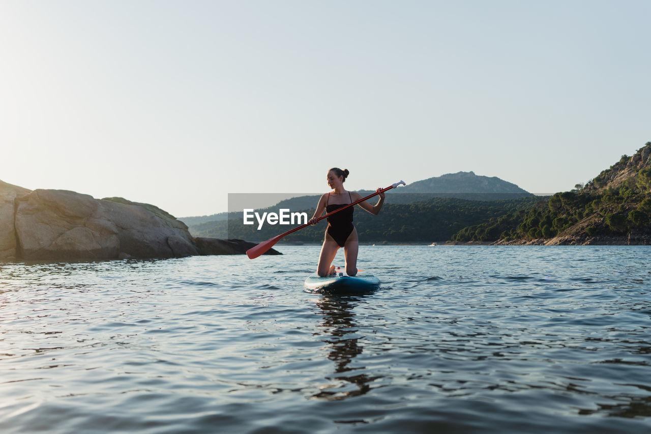 Full body of active female in swimwear floating on paddleboard in rippling lake against rock and shore with forest on summer day