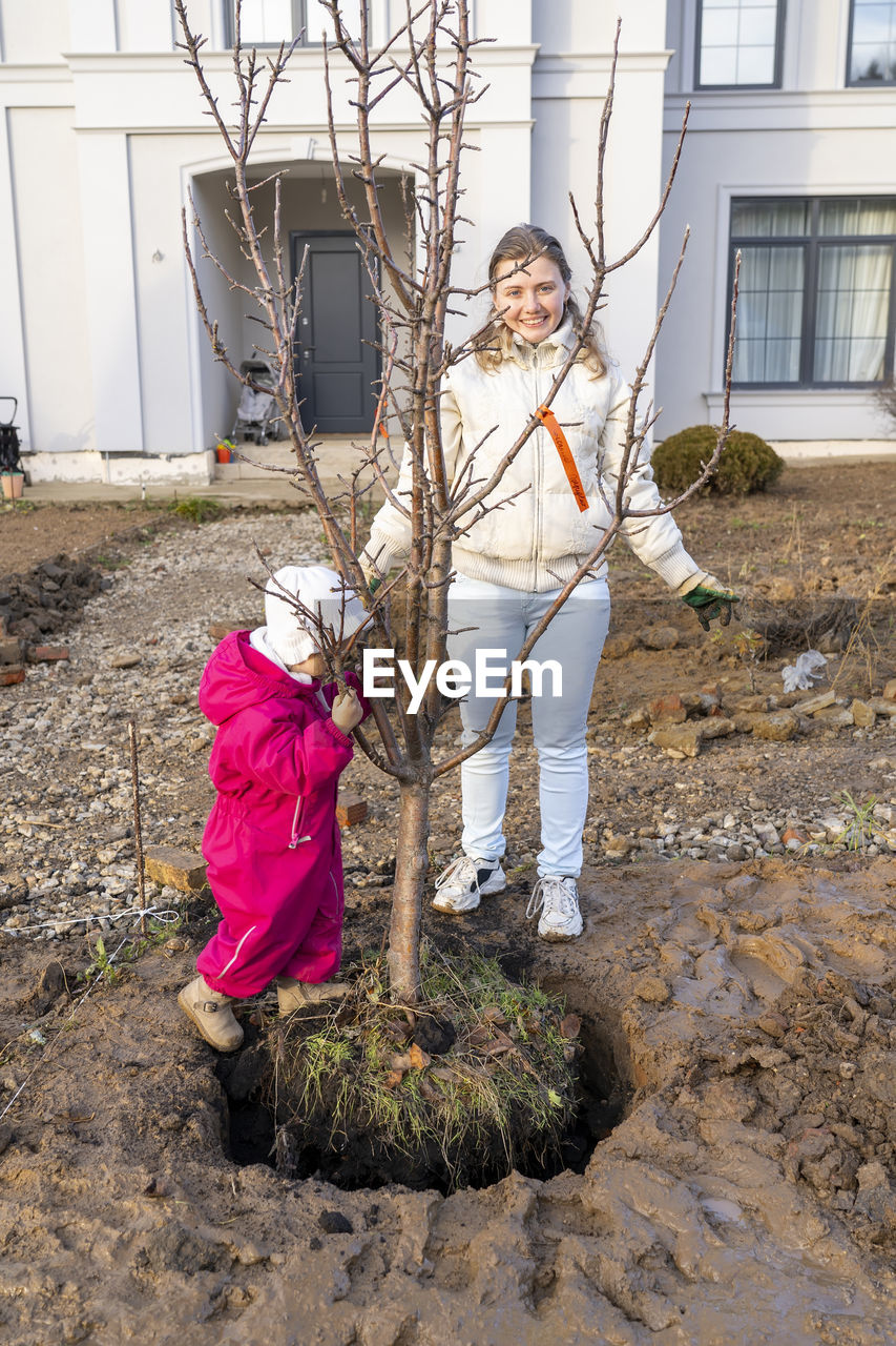 full length, child, female, childhood, women, architecture, building exterior, built structure, spring, nature, day, plant, building, flower, standing, adult, clothing, lifestyles, tree, outdoors, two people, emotion, casual clothing, looking at camera, men, family, house, front view, portrait
