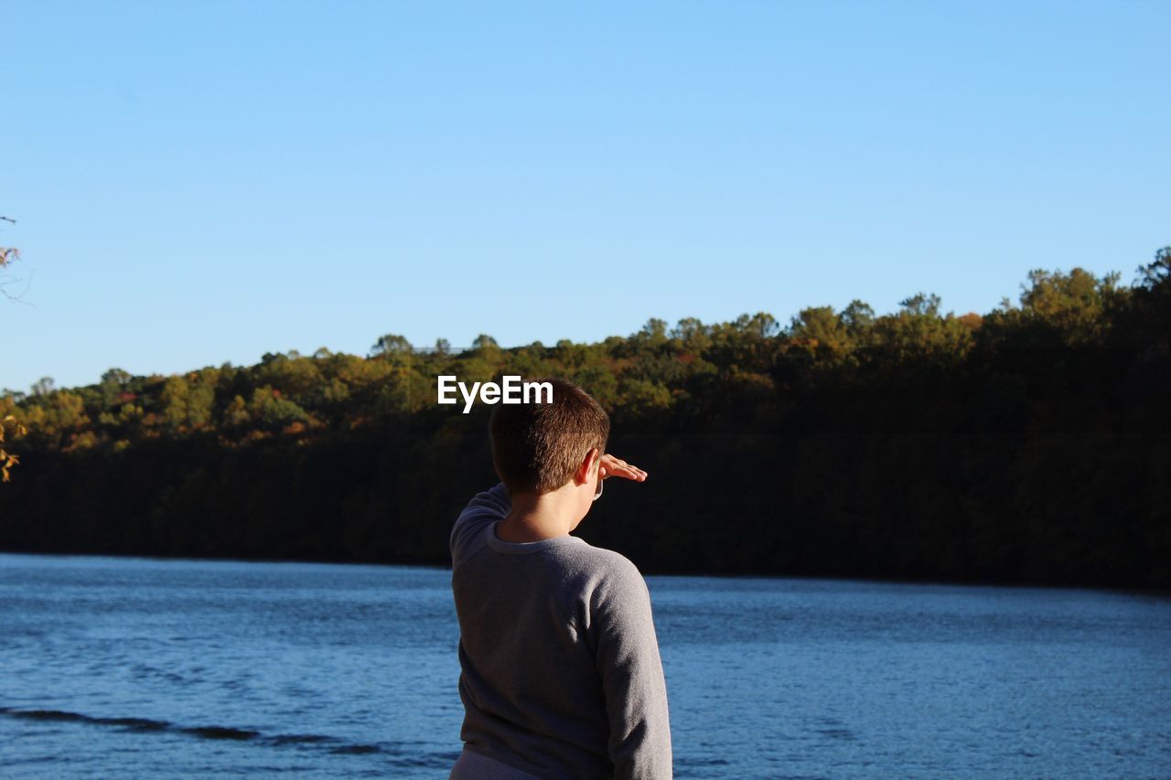 Rear view of boy shielding eyes while standing by river against clear sky