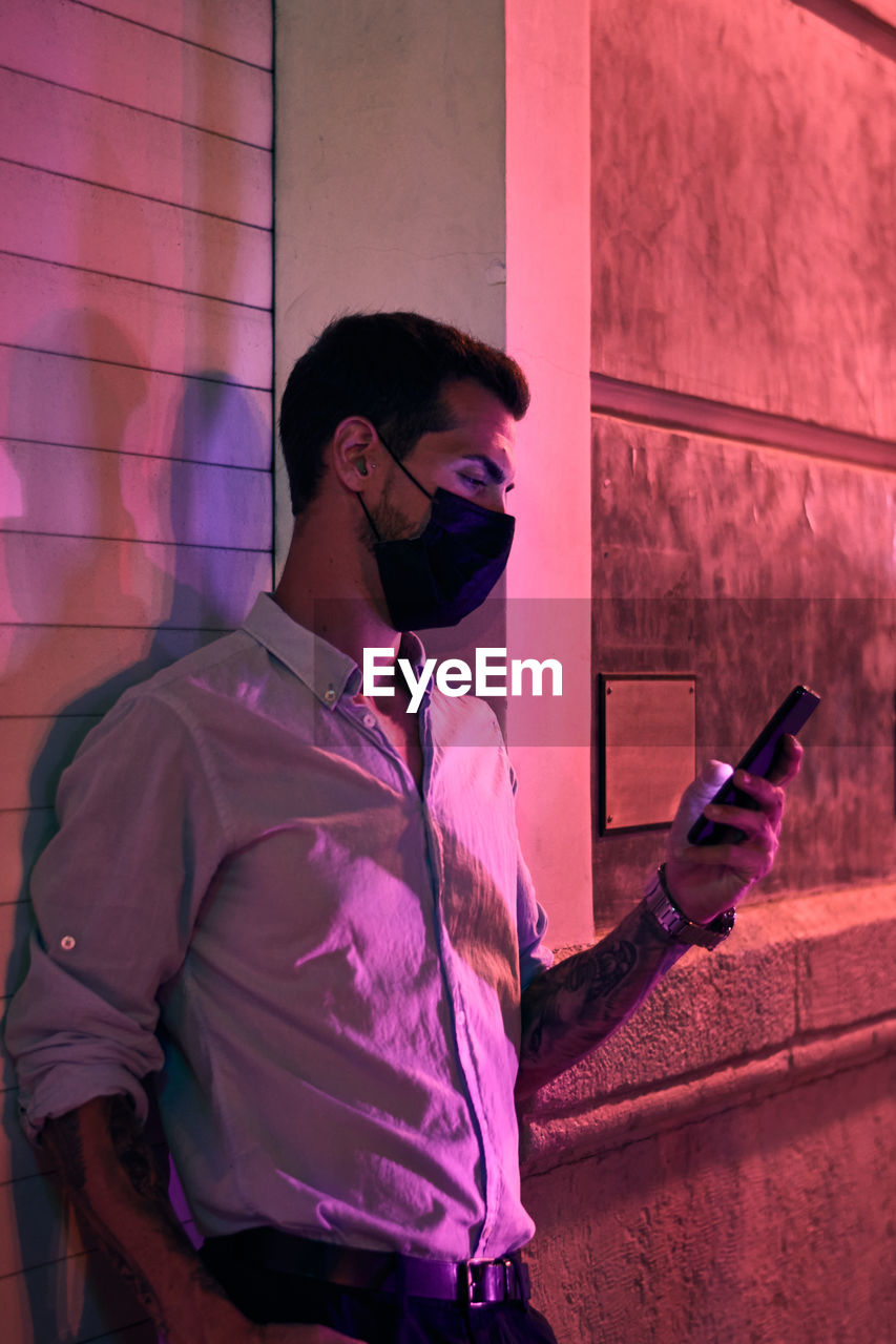 Young man with a mask looks at his mobile phone at night