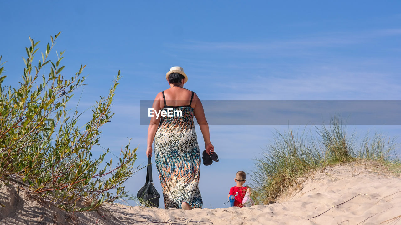 Rear view of mother and son walking at beach against blue sky