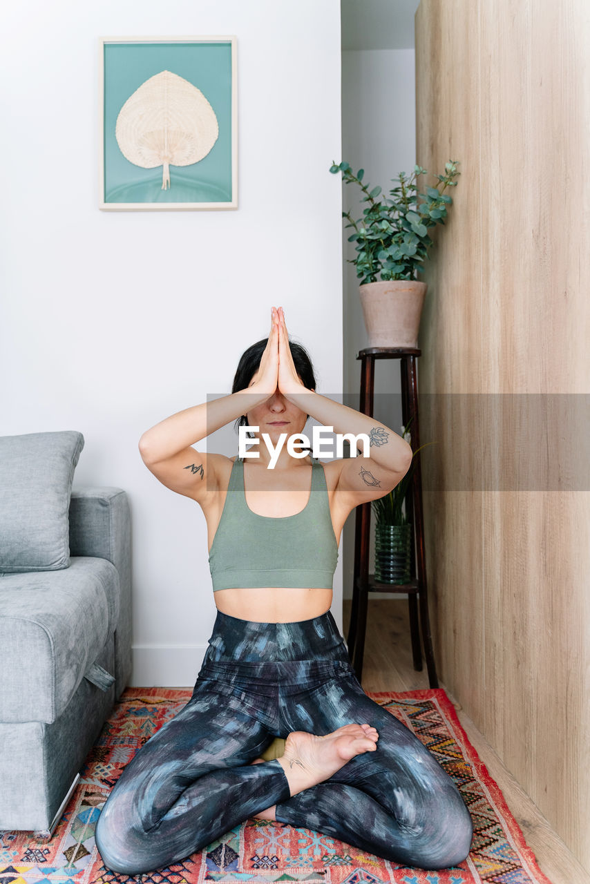 Calm hispanic female in sportswear closing eyes and doing gesturing while sitting in lotus pose on carpet near sofa during yoga session in living room at home