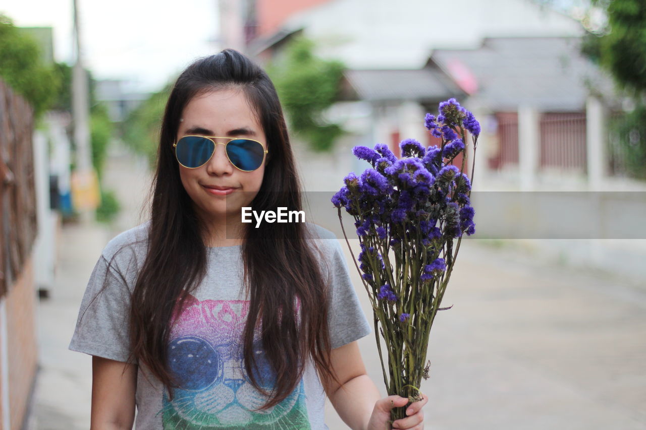 Portrait of woman wearing sunglasses while holding purple flowers on footpath