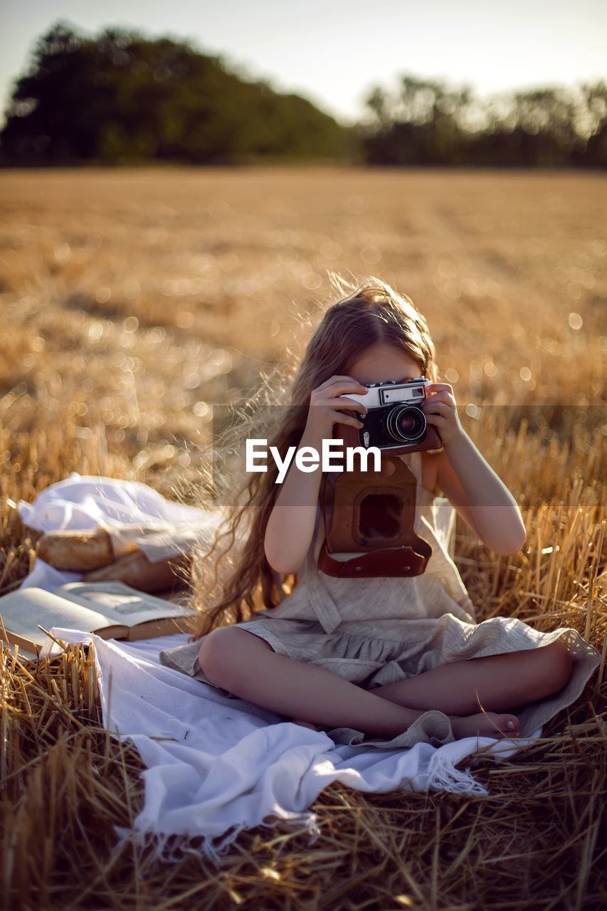 Girl child in a dress sitting on a mown field with a camera on a blanket with bread and a book