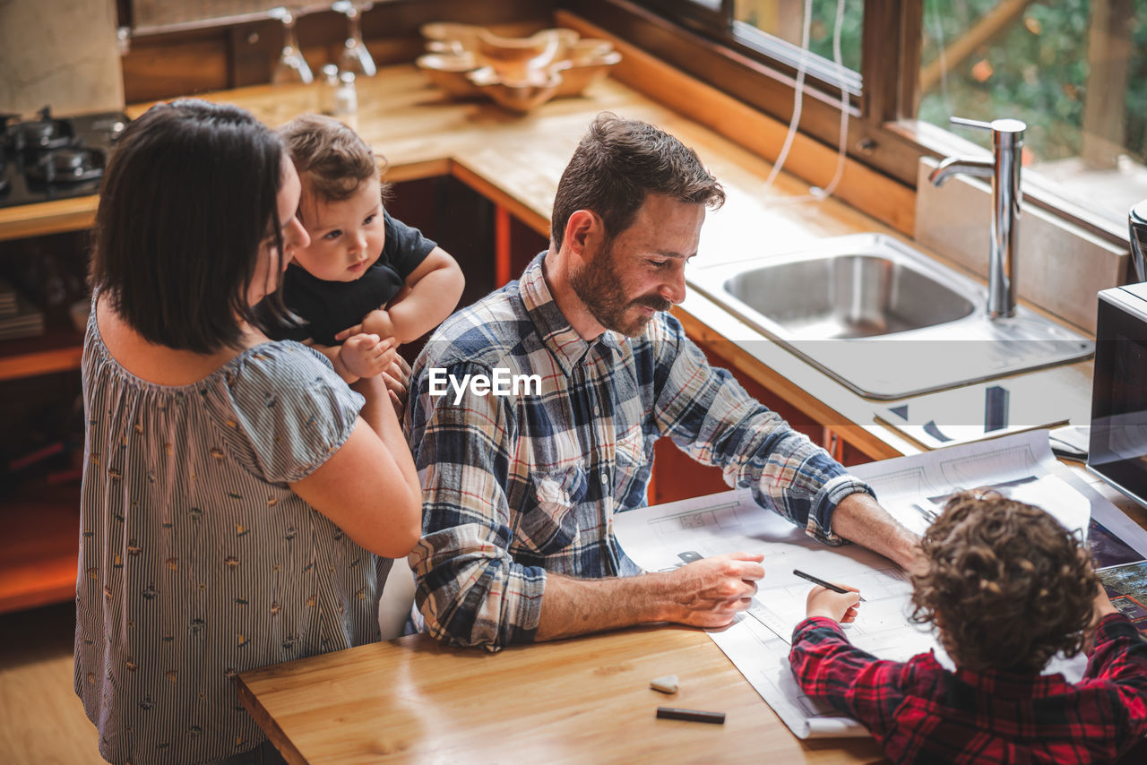 High angle of smiling male architect sitting at table and creating blueprint of house in kitchen with family