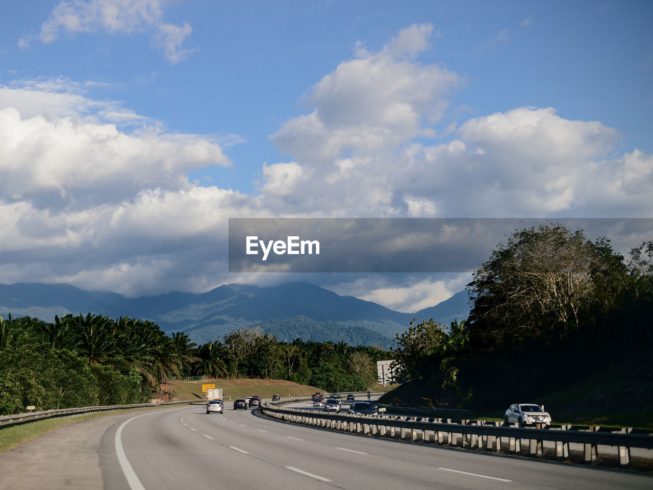 road, transportation, sky, highway, cloud, tree, nature, mountain, plant, road trip, mode of transportation, travel, freeway, symbol, infrastructure, motor vehicle, environment, the way forward, road marking, no people, landscape, car, city, marking, scenics - nature, sign, street, travel destinations, beauty in nature, horizon, land, outdoors, day, motion, traffic, asphalt, tourism, blue, journey, curve, mountain range, forest, architecture, tranquility, vanishing point, on the move