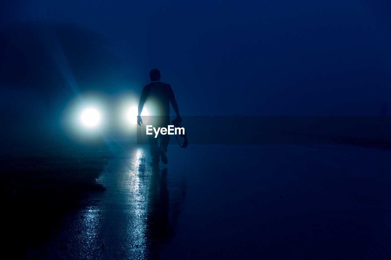 Rear view of silhouette person walking on wet street against blue sky at night