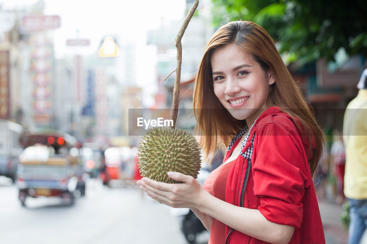 Portrait of smiling beautiful woman holding durian on street in city