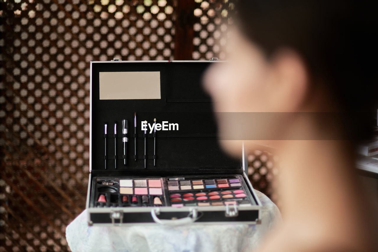 Blurred female model waiting for makeup sitting against opened suitcase with brushes and palettes on table