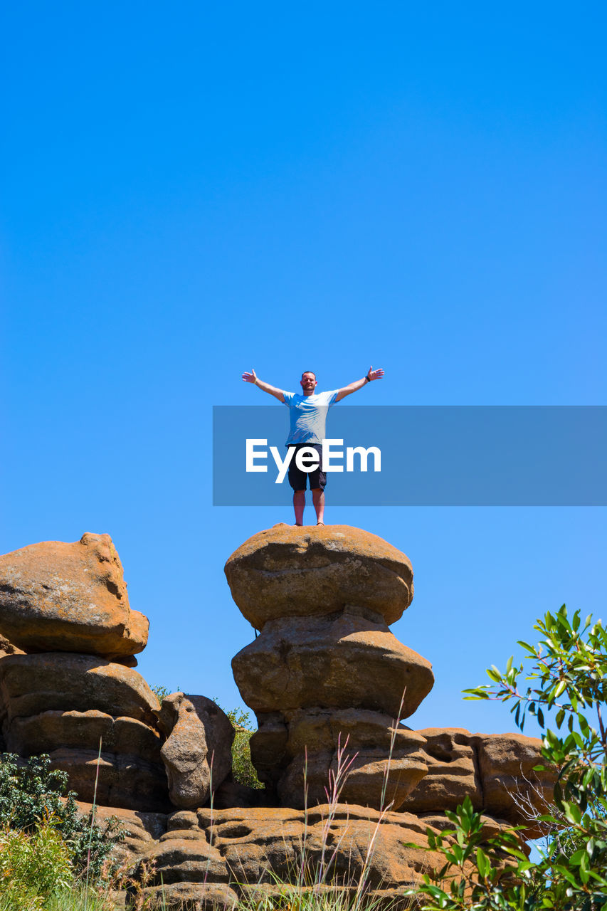 Man with arms outstretched standing on rock formation against clear blue sky