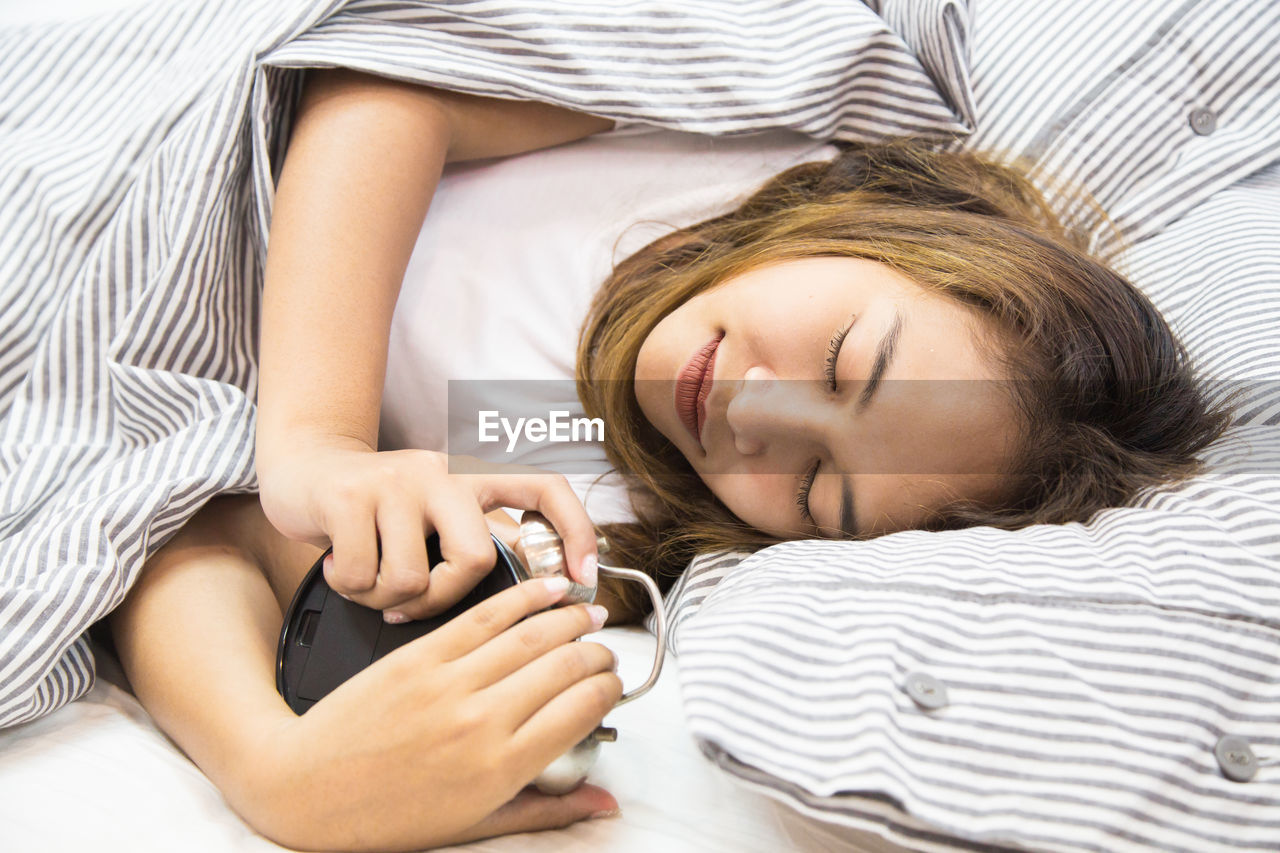 Woman holding alarm clock while sleeping on bed