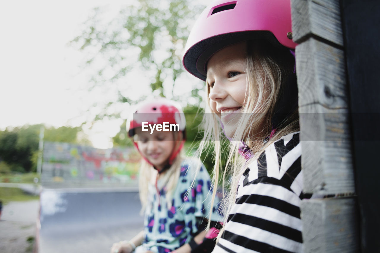 Smiling girl wearing helmet sitting with friend on edge of skateboard ramp