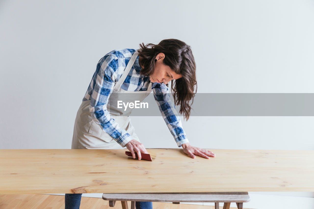 Focused skilled female woodworker with black hair in apron polishing wooden board with sandpaper while working in light professional workshop on white background