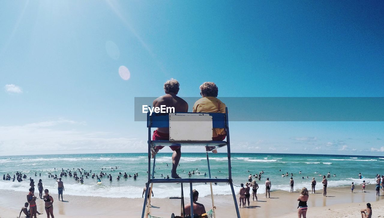 Rear view of man and woman sitting on lifeguard chair at beach against sky