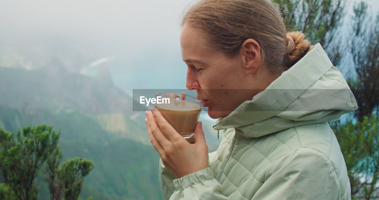 Girl enjoying cup of cappuccino in the forest at foggy morning. portrait of woman drinking coffee.