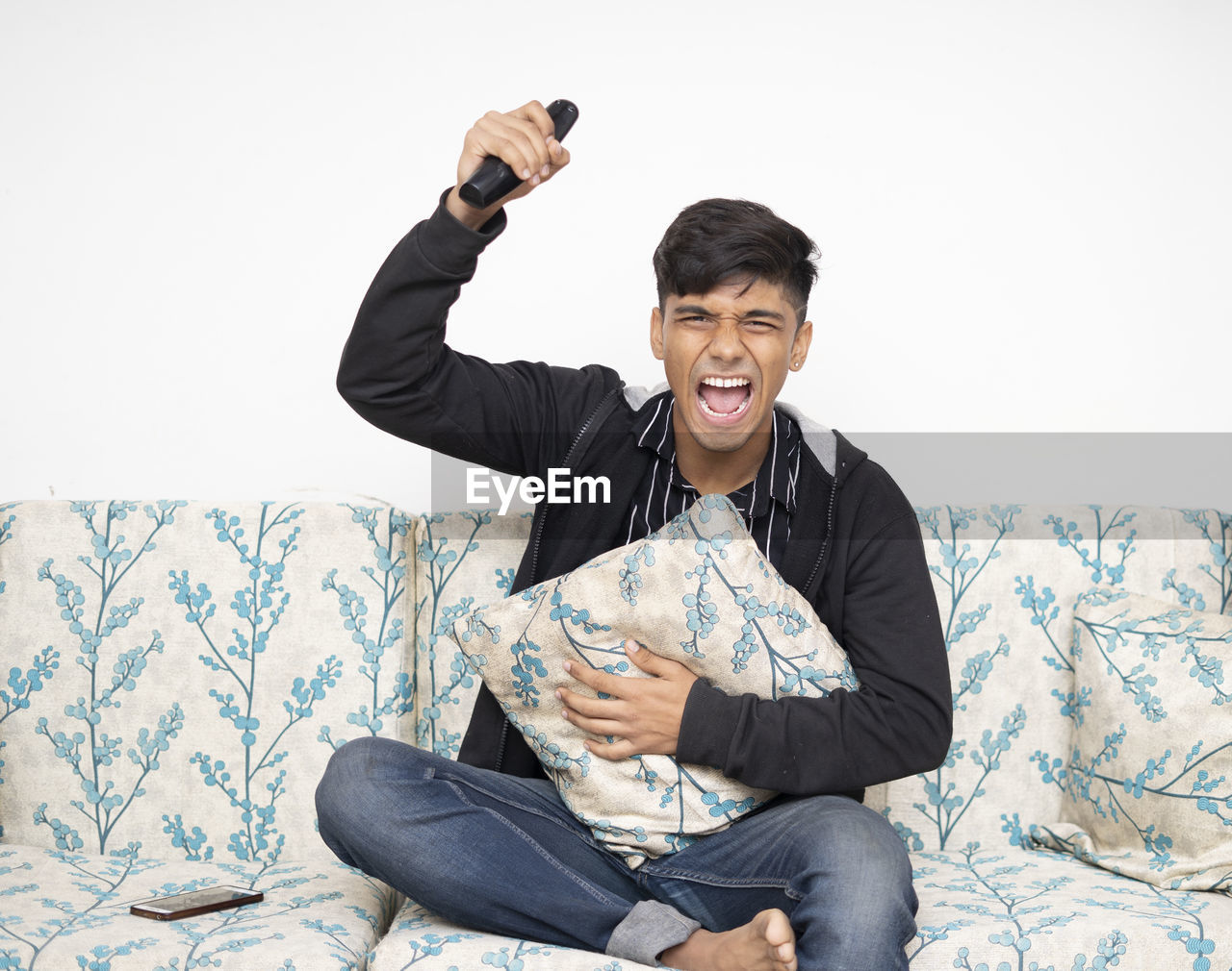 Asian man sitting on the sofa wearing black jacket holding remote while cheering for indian team.