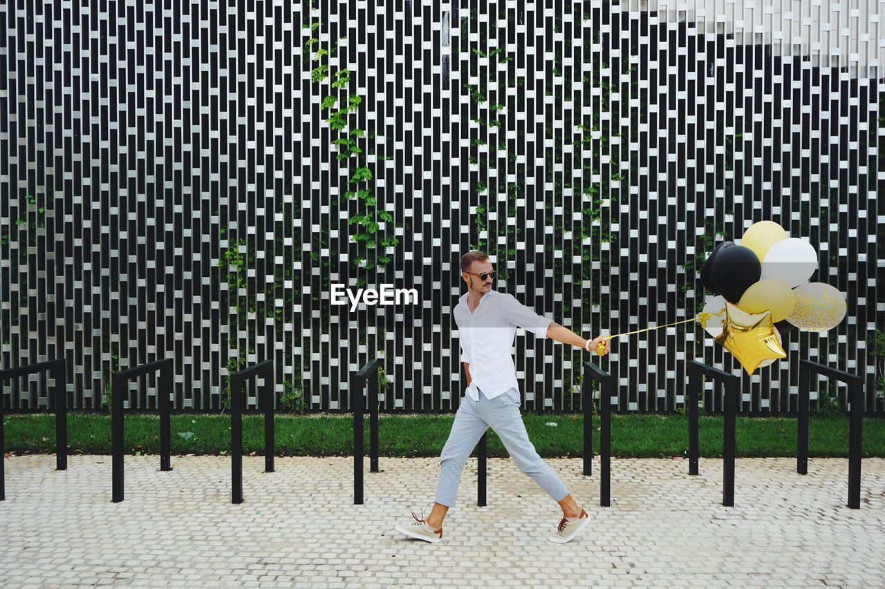 Fashionable man with balloons while walking on footpath against patterned wall