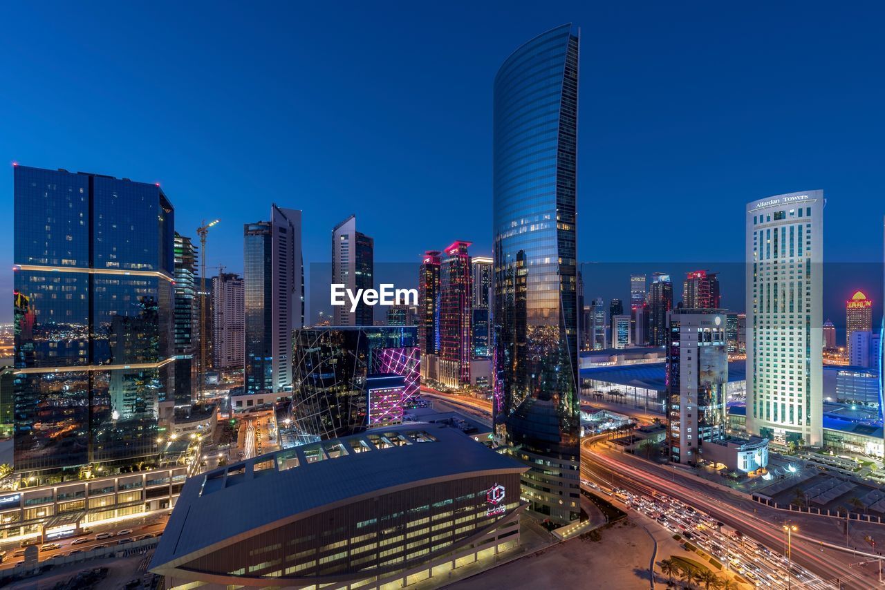 Illuminated modern buildings in city against clear blue sky