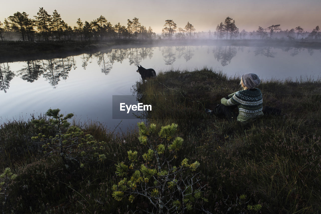 Woman and dog sit by pond at dawn with foggy atmosphere