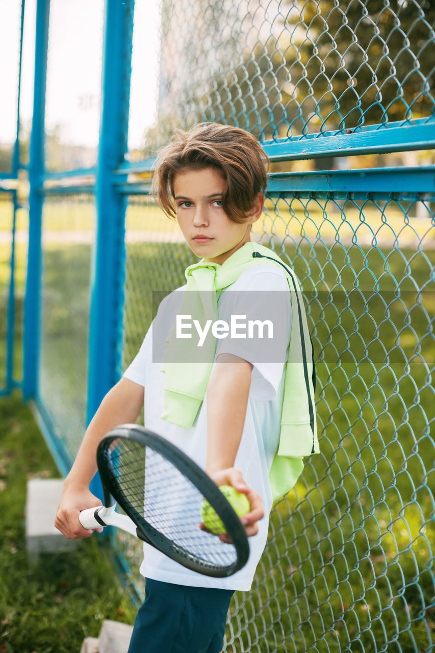 Portrait of boy with tennis racket standing by fence