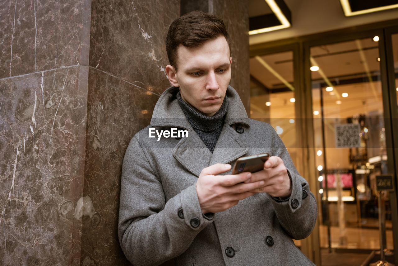 Smiling young man in gray coat is holding scroll text messages in his mobile