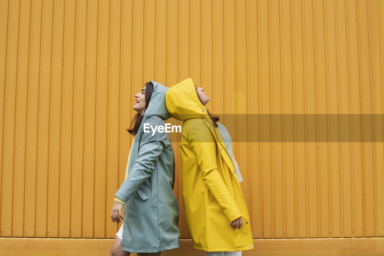 Young sister wearing blue and yellow raincoats standing back to back against wall