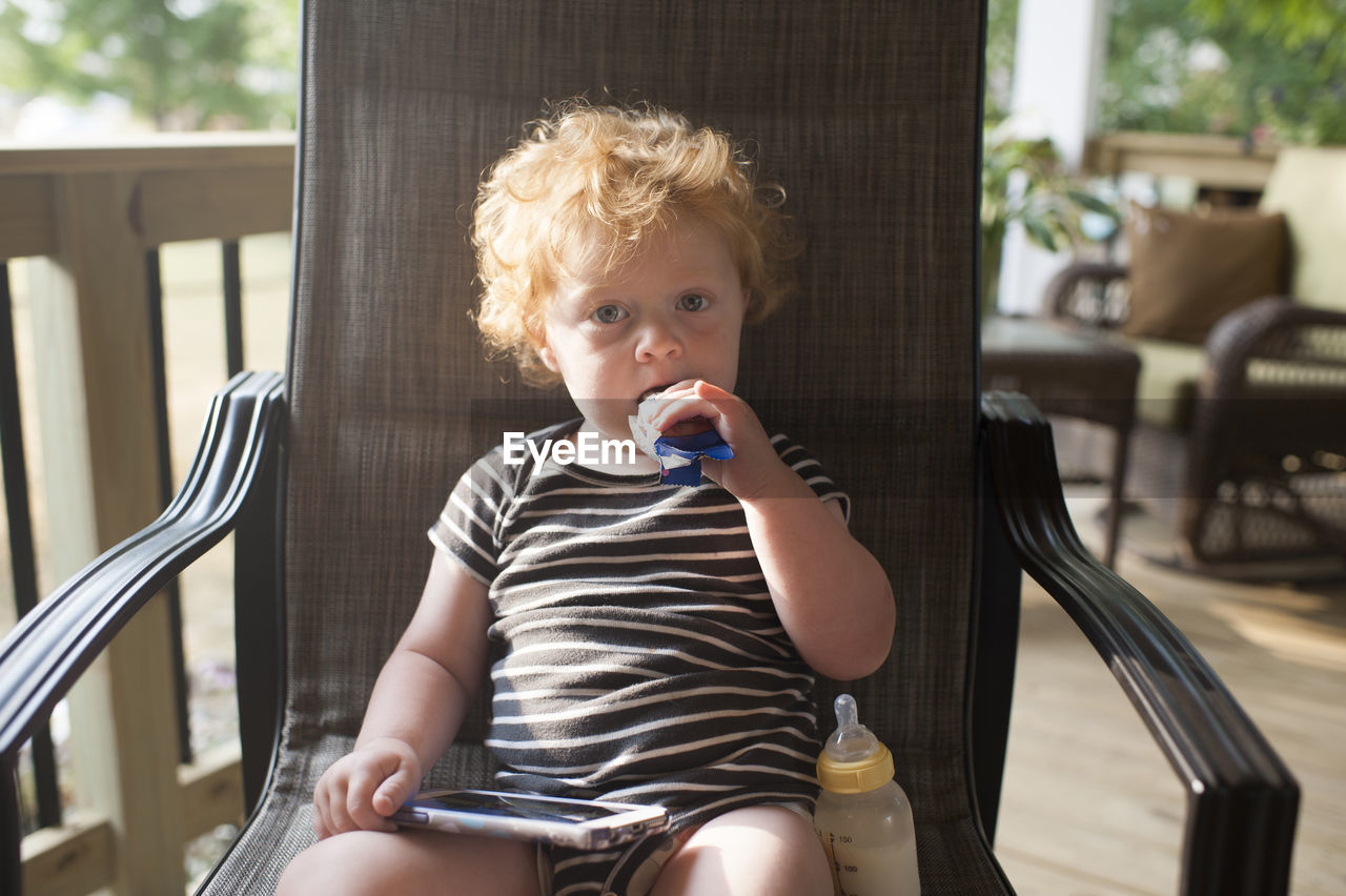 Portrait of cute baby boy eating food while sitting with smart phone and milk bottle on chair at porch