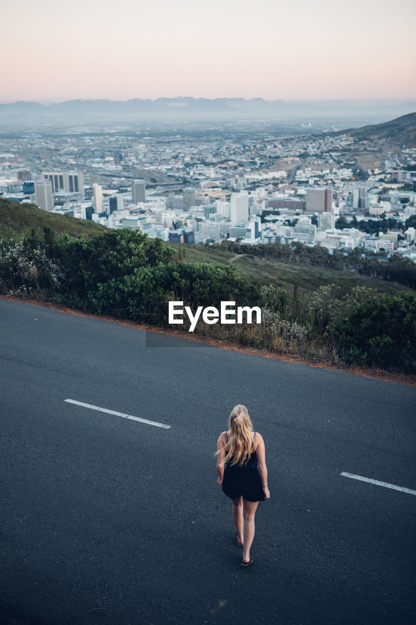 Rear view of woman standing on road by cityscape against clear sky during sunset