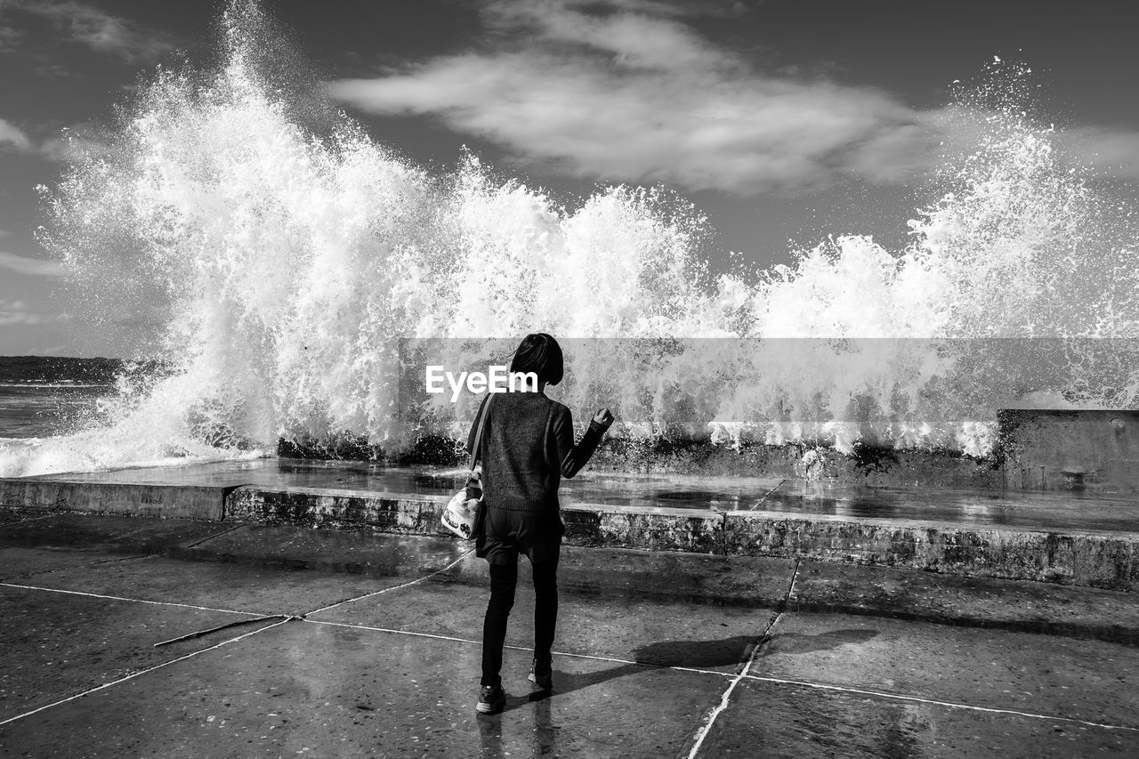 Rear view of woman standing by waves splashing at shore