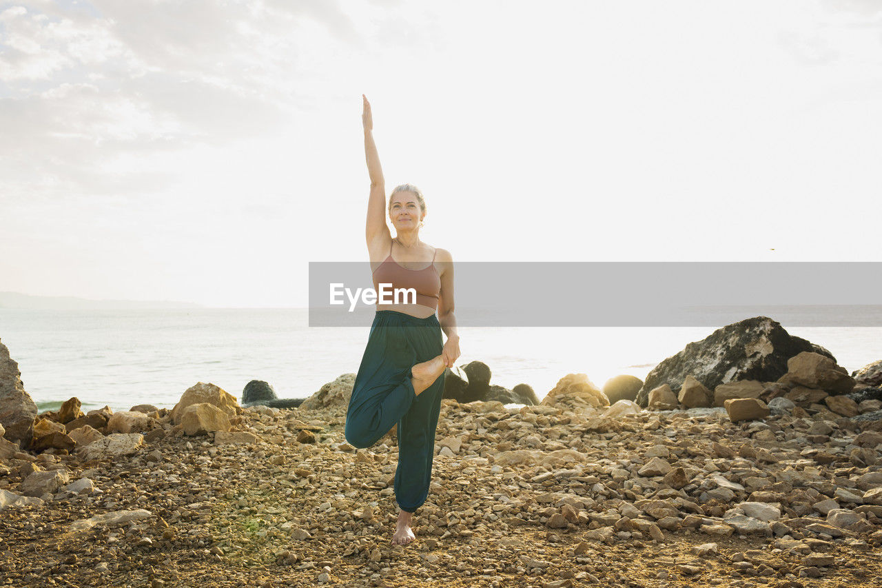 Woman practicing yoga standing on one leg in front of sky at beach
