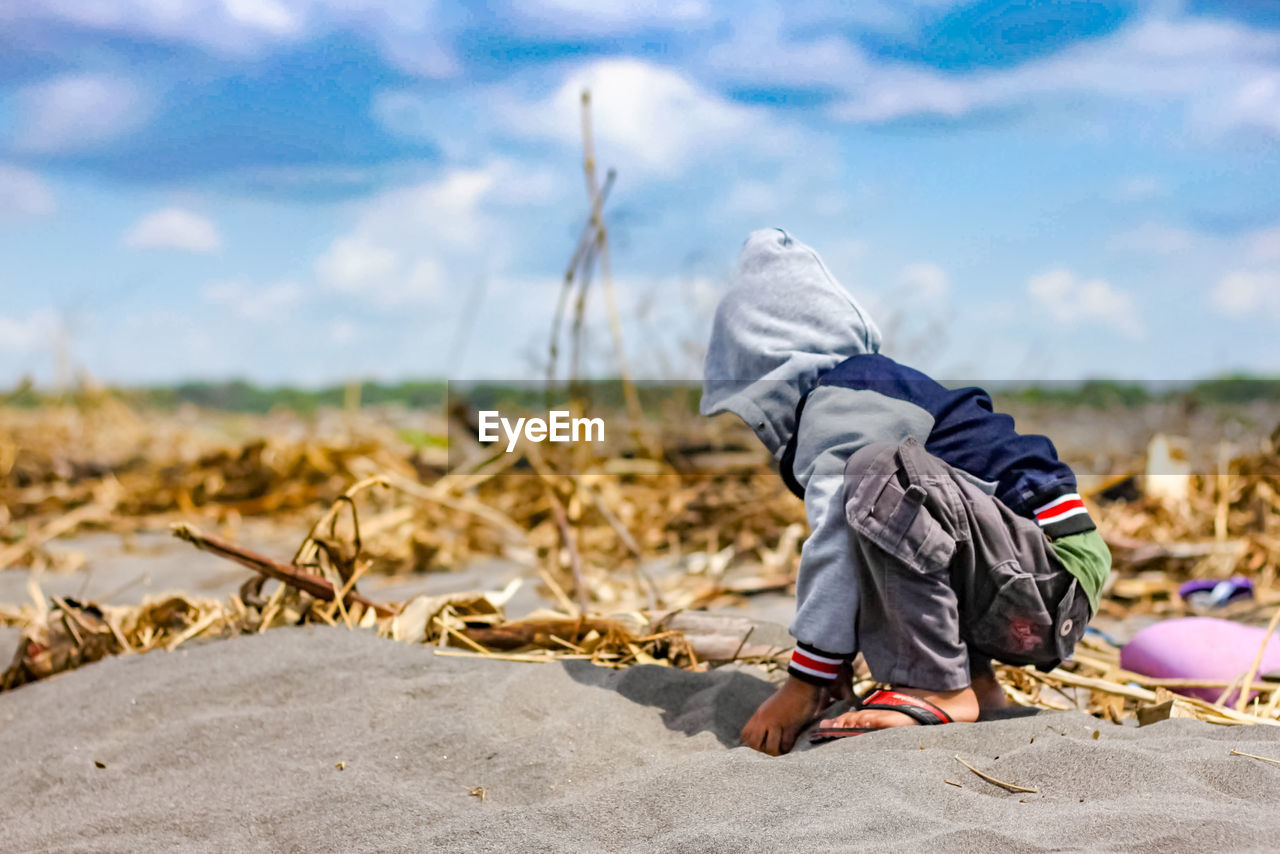 Side view of boy playing with sand at beach against sky
