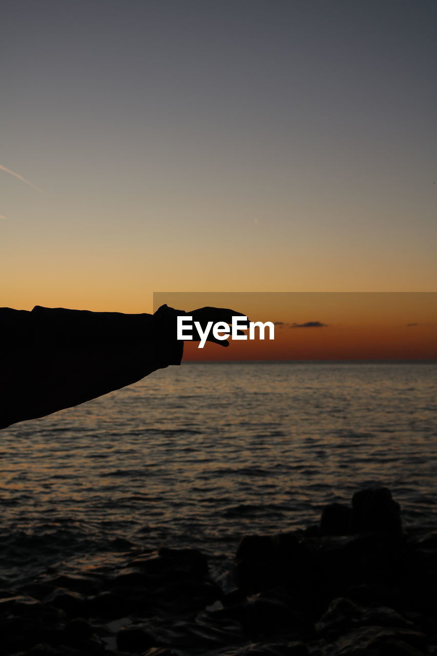 Silhouette hand by sea against sky during sunset