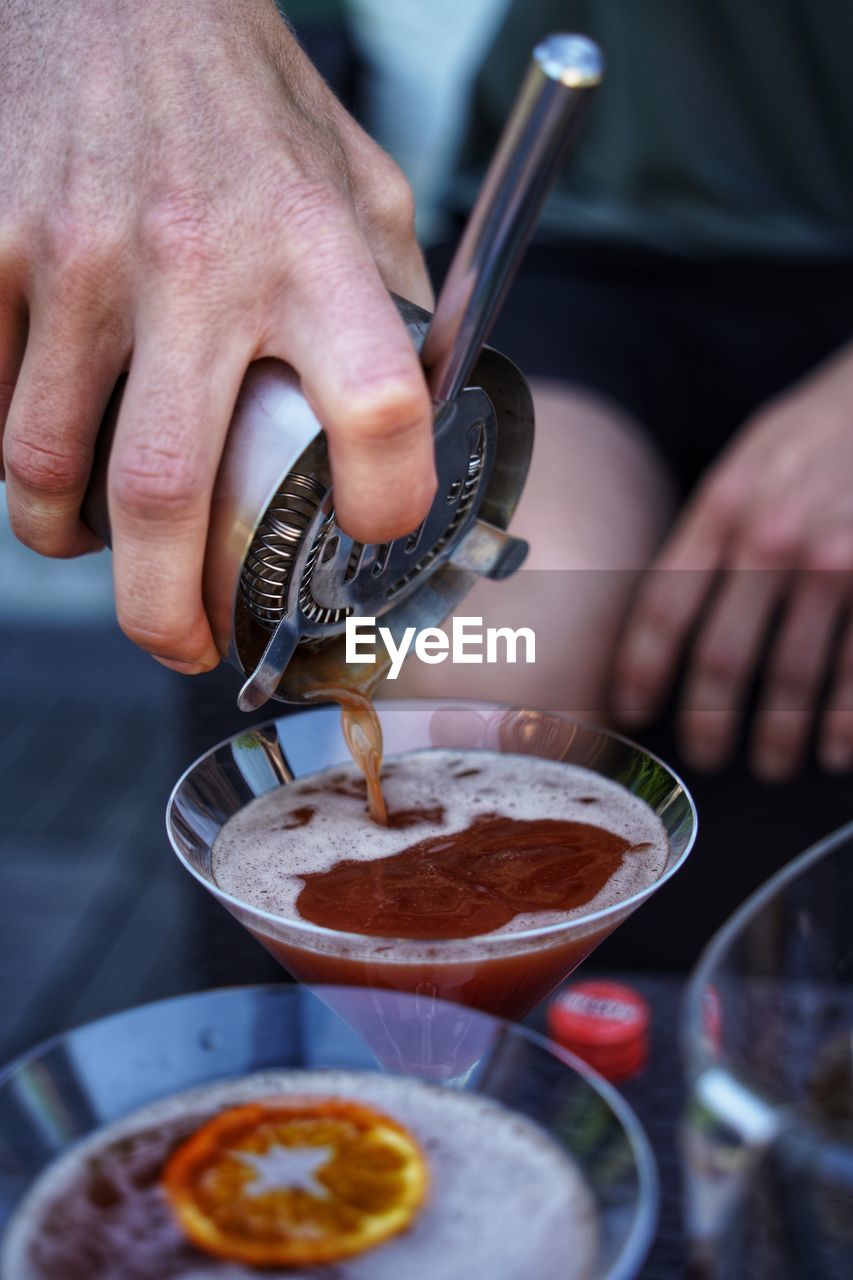 Pouring blood and sand cocktail into a martini glass with orange peel in the foreground.