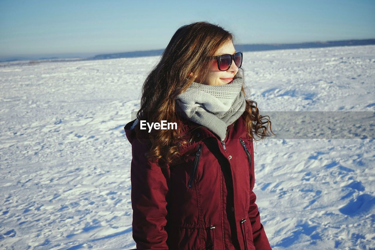 Beautiful young woman wearing muffler and jacket on snowy field against sky
