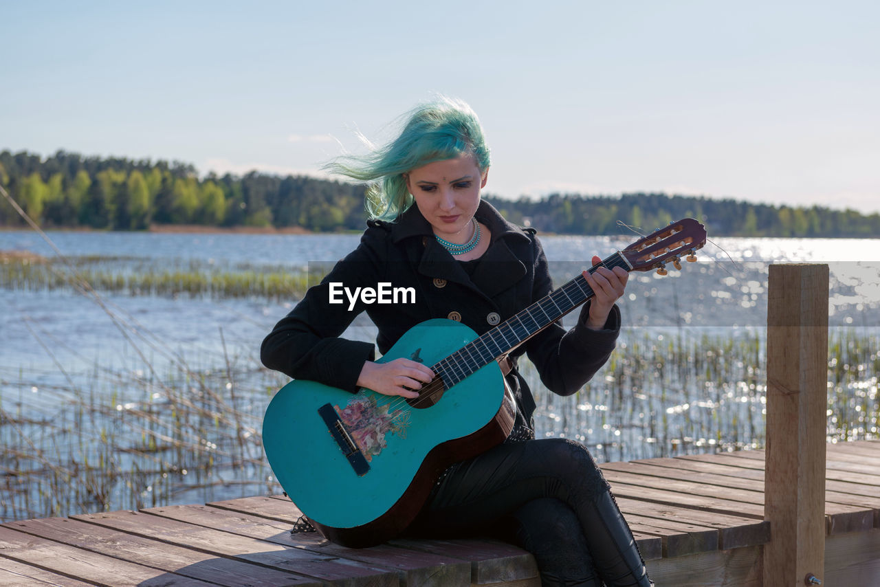 Hipster young woman with turquoise guitar sitting on pier over lake