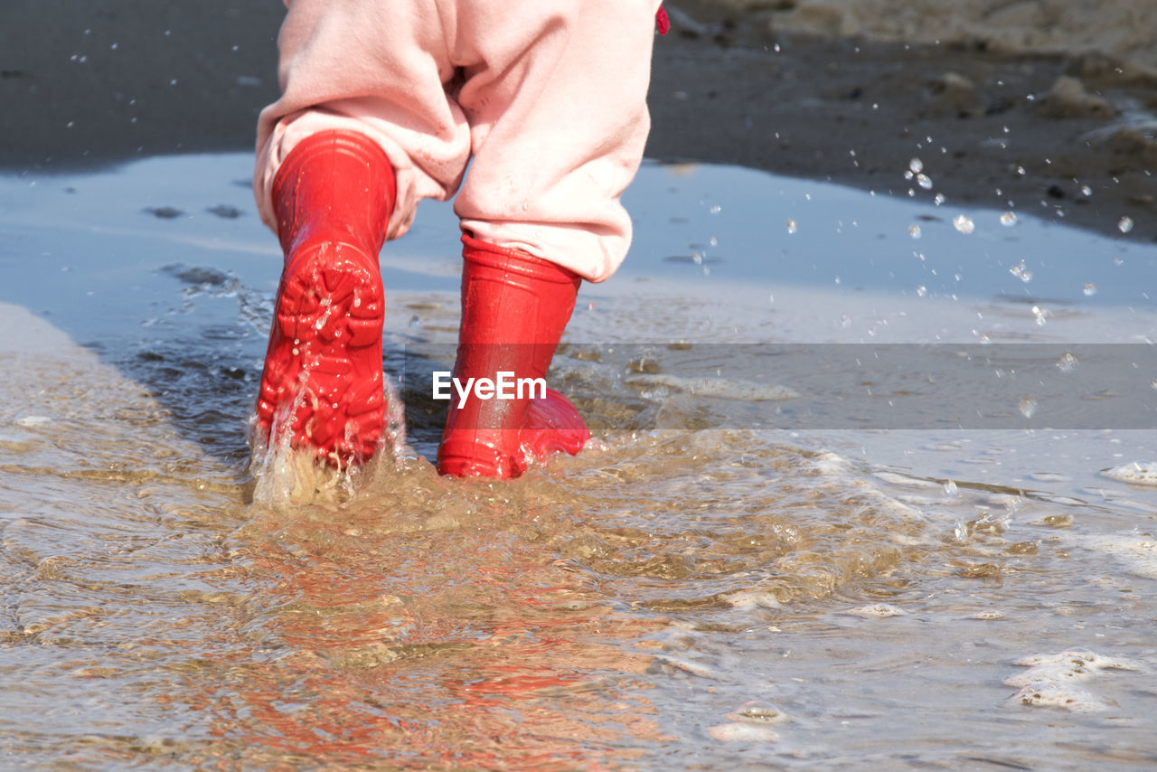 Low section of child wearing rubber boots while stamping feet in water on shore at beach