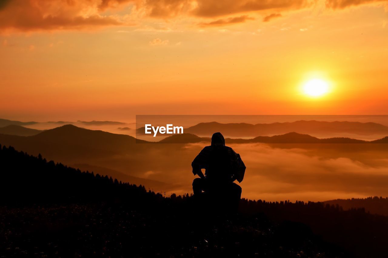 Silhouette man sitting on mountain against orange sky