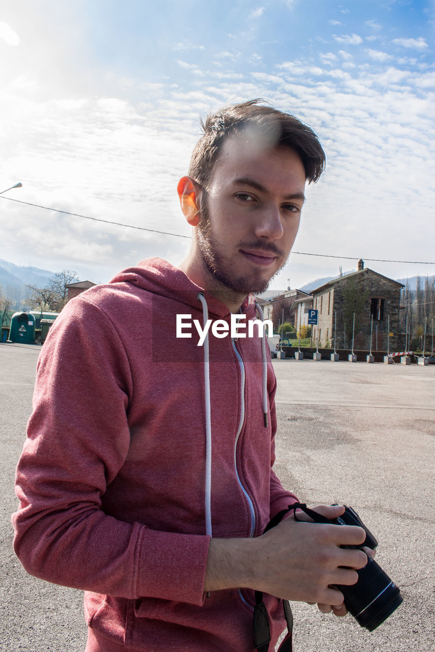 Portrait of young man holding camera while standing on street in city