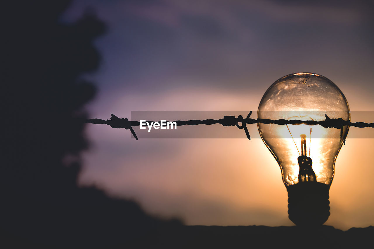 Close-up of silhouette barbed wire fence with light bulb against sky during sunset