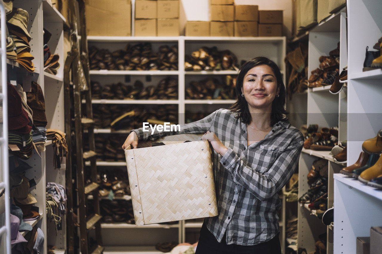 Portrait of smiling female clerk carrying wicker basket while standing amidst shoe racks at store