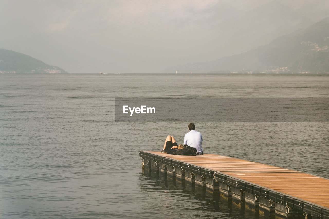 Rear view of couple of young adult sitting on the dock of the bay in lake como