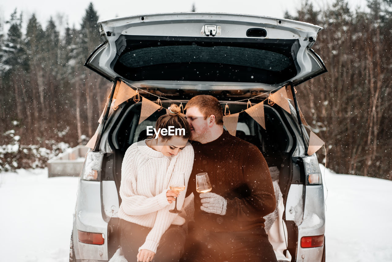 Young couple holding wineglasses standing by car during winter
