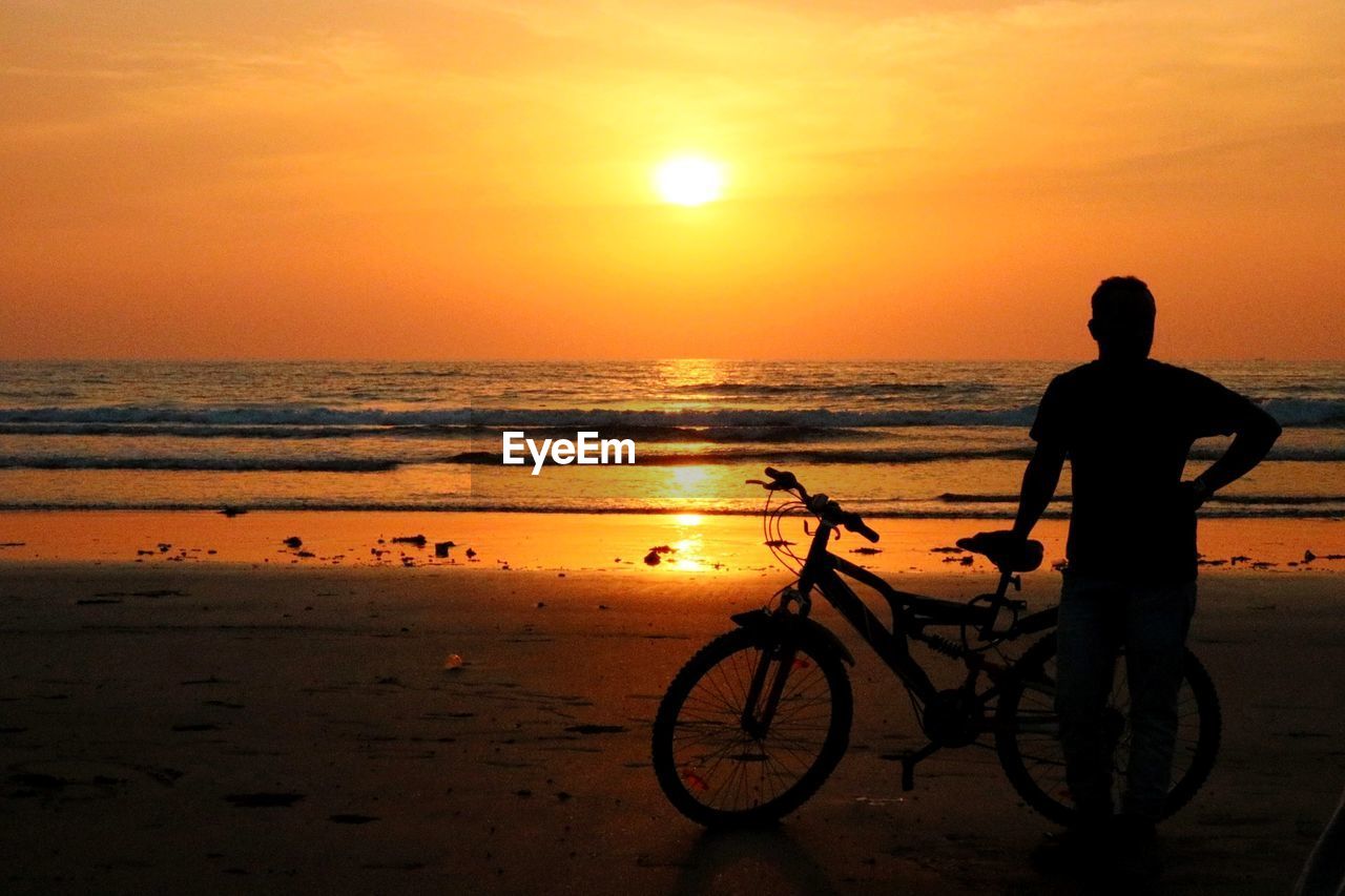 Silhouette man with bicycle standing at beach against sky during sunset